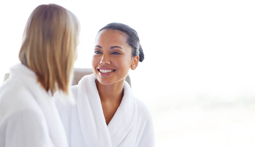 A woman dressed in a white robe smiles at another woman also dressed in a white robe.